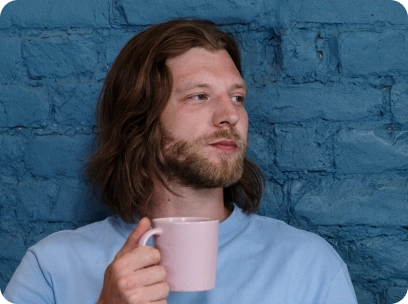 A man leans against a blue wall holding a pink coffee cup.